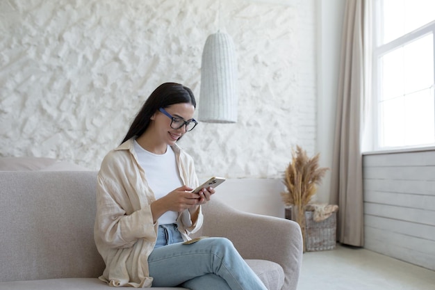 Uma jovem bonita de óculos senta-se em um sofá perto da janela e usa um telefone celular discando textos