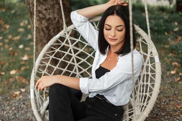 Uma jovem bonita e elegante em roupas elegantes com camisa branca senta e descansa em um balanço de malha no jardim