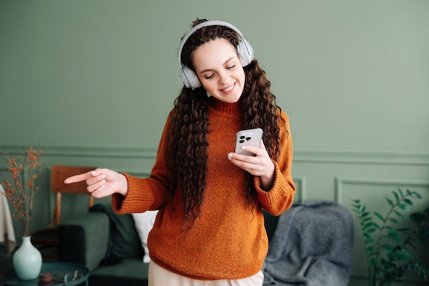 Uma jovem bonita e feliz usando fones de ouvido usando um smartphone dançando em casa, uma garota alegre