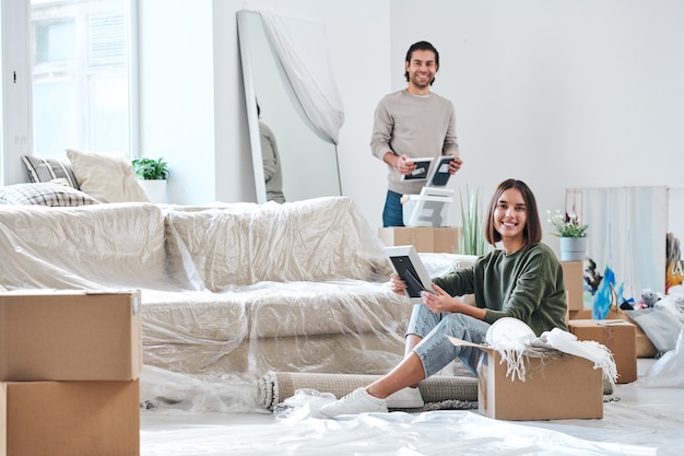 Uma jovem bonita e seu marido com porta-retratos olhando para você enquanto desempacota caixas em sua nova casa