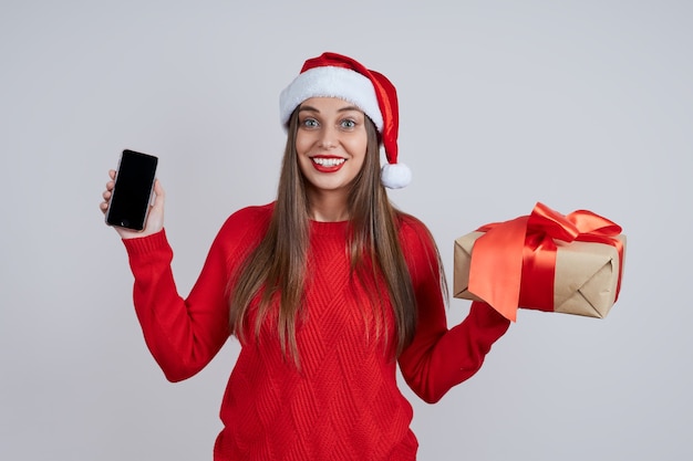 Uma jovem bonita e sorridente com um chapéu de Papai Noel, com um presente e um telefone nas mãos. Conceito de compras online, saudações online, descontos de Natal.