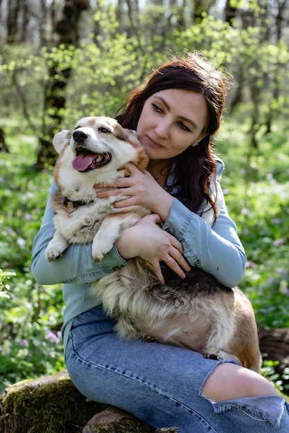uma jovem bonita em jeans e uma camisa jeans abraça suavemente um cachorro corgi na floresta de primavera
