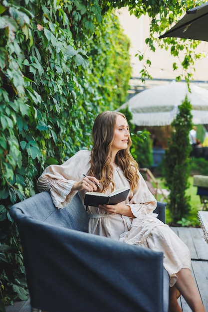 Uma jovem bonita em um vestido senta-se com um caderno e faz anotações no terraço de um café de verão Mulher em roupas de verão casuais e elegantes