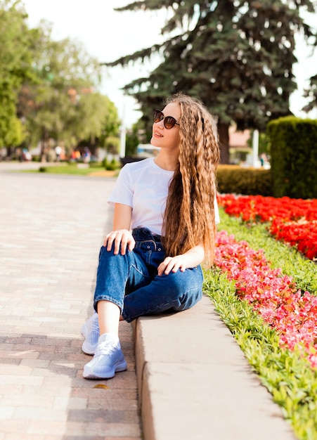 Uma jovem bonita em uma camiseta branca e jeans em óculos de sol senta-se perto de um canteiro de flores