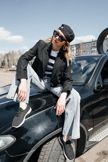 Uma jovem bonita está sentada no capô de um carro. Uma garota elegante de terno e óculos em um carro preto.