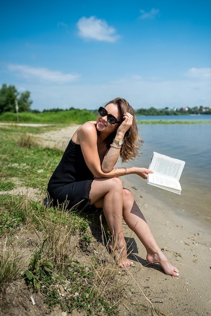 Uma jovem bonita no dia de verão leu o livro perto de um estilo de vida do lago