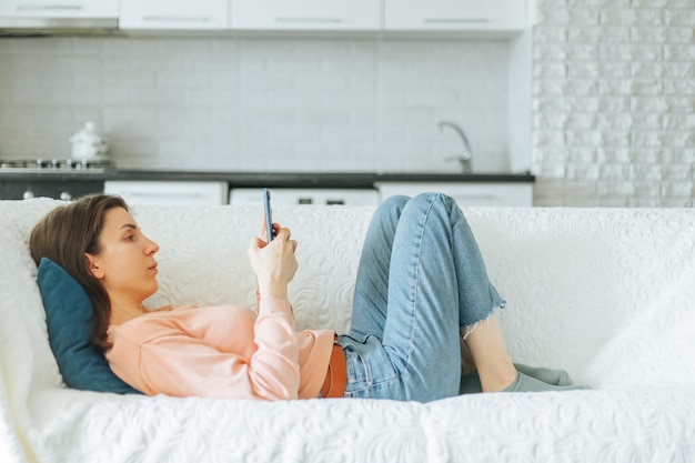 Uma jovem bonita senta-se confortavelmente em seu sofá em casa, absorta na leitura de seu smartphone.