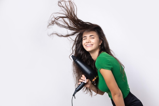 Uma jovem bonita vestida com um top verde seca seus belos cabelos longos e sedosos com um secador de cabelo