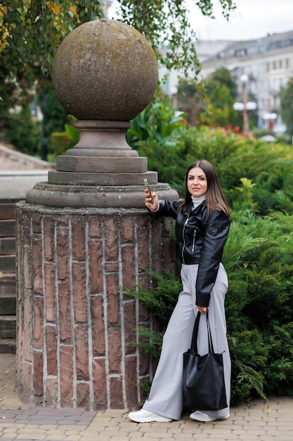 Uma jovem bonita vestindo uma jaqueta de couro preta caminha pela rua em um parque e usa telefone