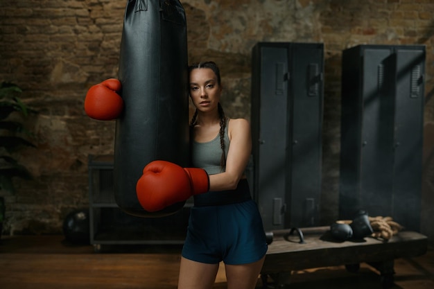 Uma jovem boxeadora confiante usando luvas descansa perto do saco de pancadas no antigo ginásio esportivo