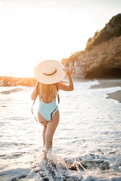 Uma jovem bronzeada em um lindo maiô com chapéu de palha se levanta e descansa em uma praia tropical com areia e olha o pôr do sol e o mar. Foco seletivo. Conceito de férias à beira-mar