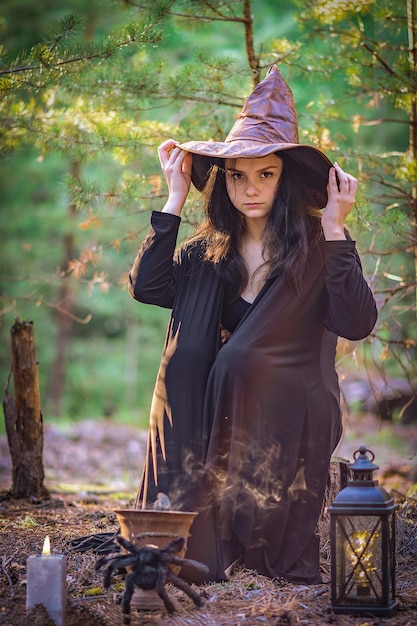 Foto uma jovem bruxa na floresta está sentada em um toco, segurando seu chapéu.