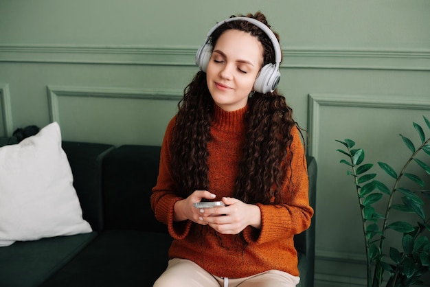 Uma jovem calma e despreocupada ouve música em um podcast de áudio ou audiolivro em seu telefone celular enquanto está sentada em casa meditando e relaxando sentindo paz de espírito com os olhos fechados