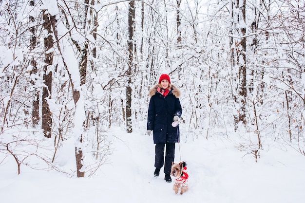 Uma jovem caminha na floresta inferior no inverno com um cachorro vestindo um suéter de natal