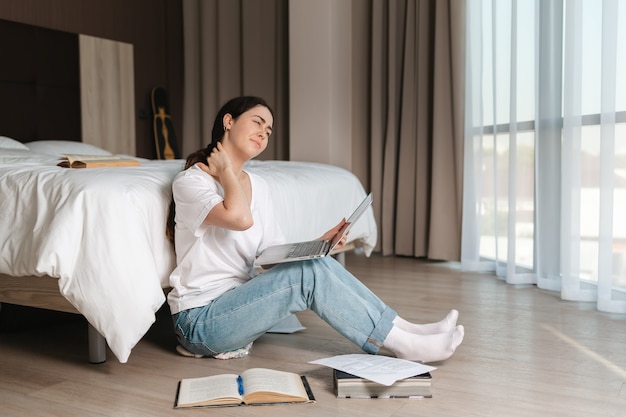 Uma jovem cansada faz uma massagem no pescoço enquanto trabalhava em seu laptop. Interior da casa. O conceito de aprendizagem e trabalho online.