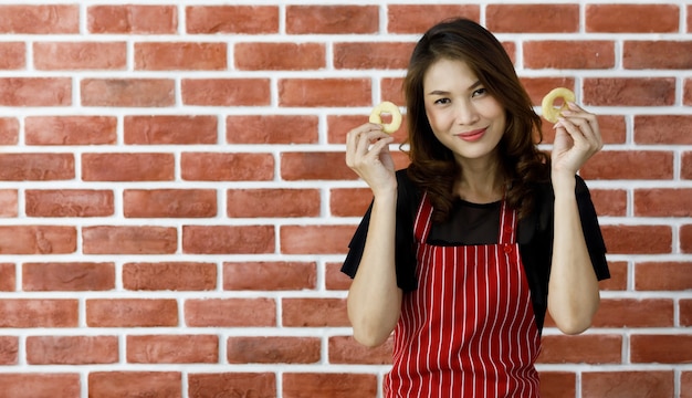 Uma jovem chef asiática com avental listrado vermelho fica perto da parede de tijolos e brinca para mostrar o anel de cebola com um sorriso antes de fritar na frigideira preta na mesa de utensílios de cozinha e ingredientes