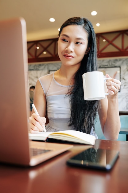 Uma jovem chinesa bebendo uma caneca de café enquanto trabalha em um laptop em um café local, depois das aulas na faculdade