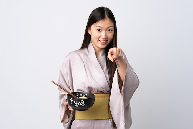 Uma jovem chinesa vestindo quimono sobre um fundo branco isolado aponta o dedo para você com uma expressão confiante, segurando uma tigela de macarrão com pauzinhos