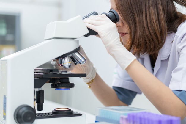 Uma jovem cientista com um jaleco olhando através de um microscópio em um laboratório para fazer pesquisas e experimentos. Cientista trabalhando em um laboratório. Foto de estoque de educação