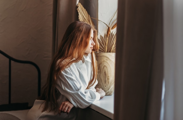 Uma jovem com cabelo comprido sentada na cama depois de acordar olhando para a cidade pela janela, dentro de casa em casa de manhã cedo, estilo de vida