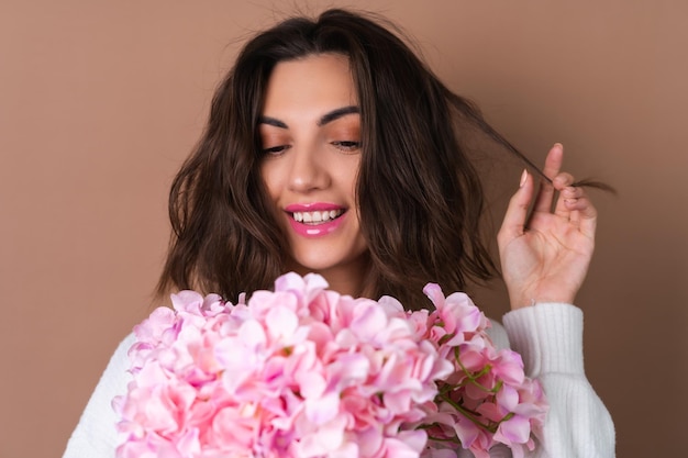 Uma jovem com cabelo volumoso ondulado em um fundo bege com brilho labial de batom rosa brilhante em um suéter branco segura um buquê de flores cor de rosa