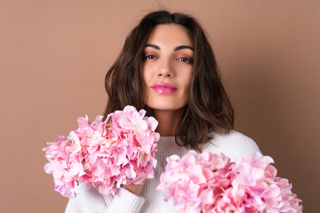 Uma jovem com cabelo volumoso ondulado em um fundo bege com brilho labial de batom rosa brilhante em um suéter branco segura um buquê de flores cor de rosa