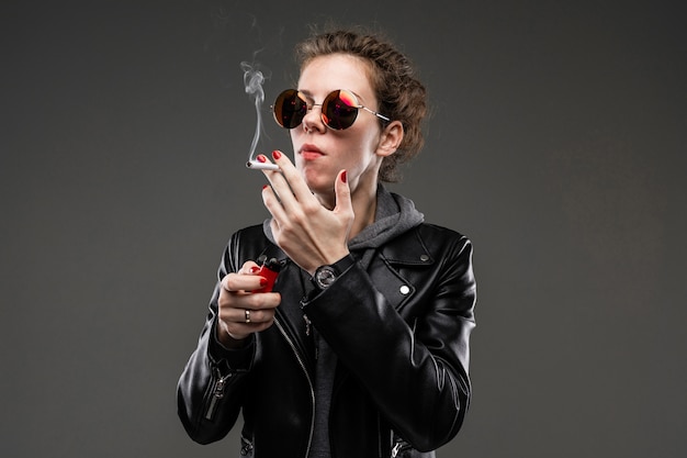 Uma jovem com características faciais ásperas em uma jaqueta preta fuma isoladas no fundo preto