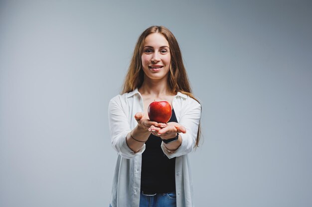 Uma jovem com longos cabelos morenos vestidos com uma camisa branca e jeans tem uma maçã vermelha fresca nas mãos conceito de comida saudável fundo branco