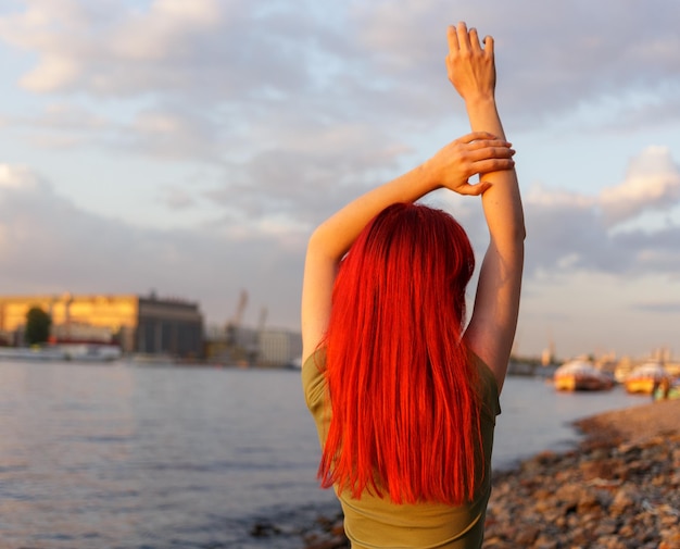 Uma jovem com o cabelo vermelho solto com as mãos levantadas posa para a câmera nos raios do pôr-do-sol
