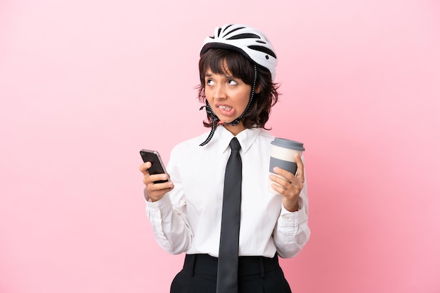 Uma jovem com um capacete de bicicleta isolado em um fundo rosa segurando um café para levar e um celular enquanto pensa em algo