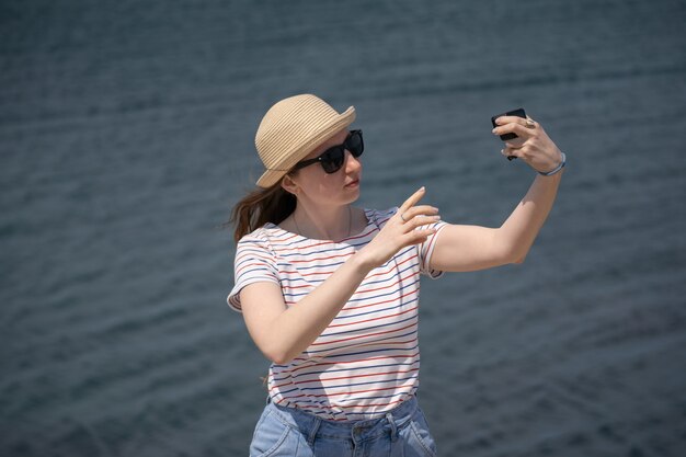 Uma jovem com um chapéu de palha e óculos escuros faz uma selfie usando uma câmera de ação no lago touris ...