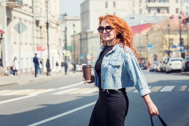 Uma jovem com um copo de café caminha na cidade em um dia ensolarado e quente Beleza ruiva em óculos de sol e uma jaqueta jeans