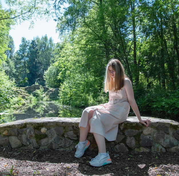 Uma jovem com um olhar surpreso em um vestido rosa está sentada em uma ponte de pedra na floresta em um dia ensolarado de verão