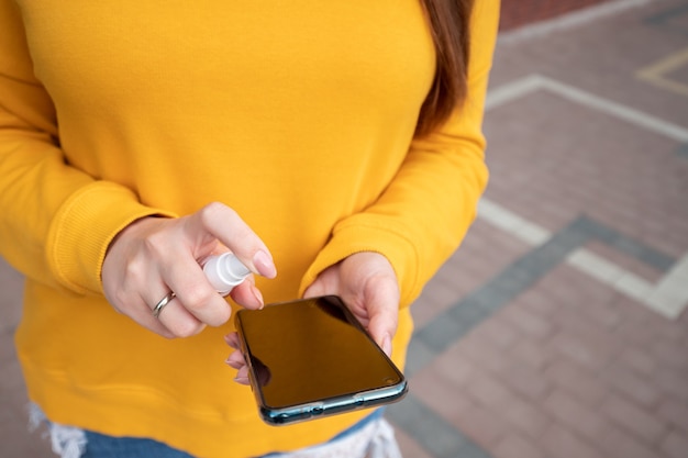Uma jovem com um suéter amarelo desinfeta o telefone com um spray desinfetante