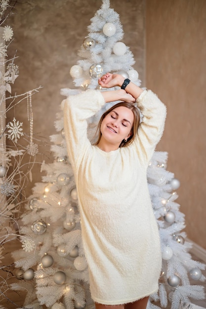 Uma jovem com um suéter branco em um quarto decorado para o Natal está de pé contra o fundo de uma árvore de Natal