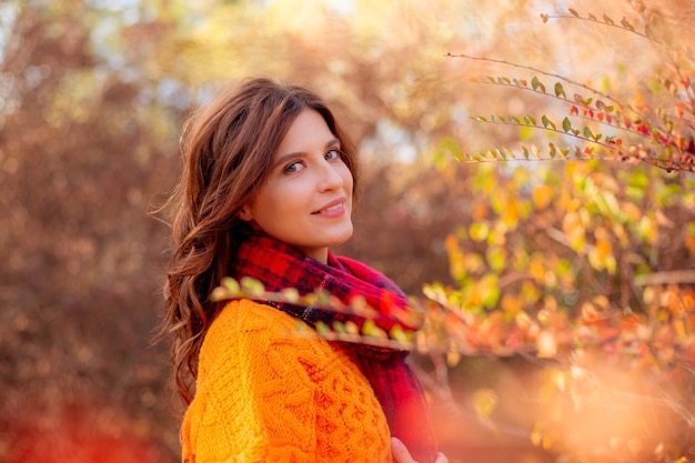 Uma jovem com um suéter laranja se envolve em um lenço em um parque de outono
