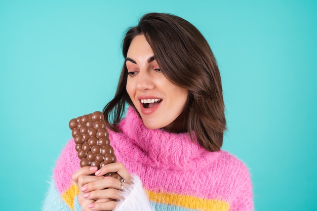 Uma jovem com um suéter multicolorido brilhante em azul olha com desejo em uma deliciosa barra de chocolate ao leite arejado