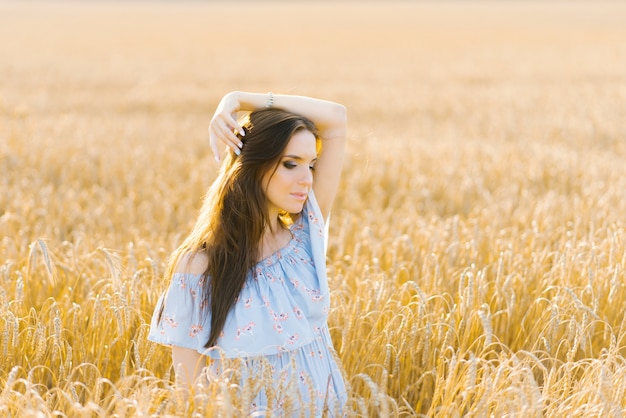 Uma jovem com um vestido azul parada em um campo de centeio ao pôr do sol