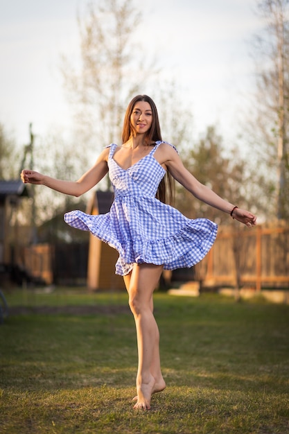Foto uma jovem com um vestido curto xadrez está dançando na grama verde do parque em um dia quente. o conceito de férias de verão na vila e o estilo de vida