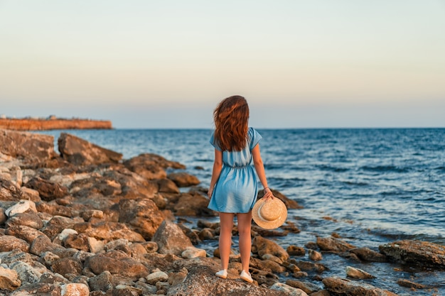 Uma jovem com um vestido e um chapéu em pé na praia ao pôr do sol e olhando para o sol poente