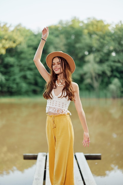 Uma jovem com um visual hippie e ecodress viaja na natureza à beira do lago usando um chapéu e calças amarelas no pôr do sol de outono