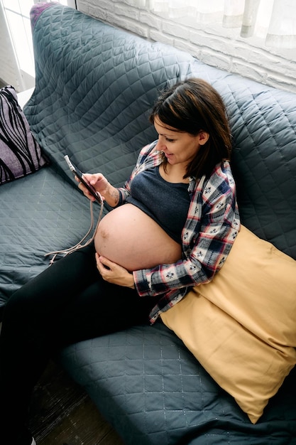 Uma jovem com uma gravidez muito avançada está sentada no sofá olhando e falando em seu celular