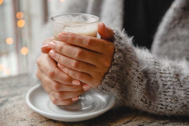 Uma jovem com uma jaqueta quente está sentada em um café à mesa perto da janela e aquece as mãos em um copo de café