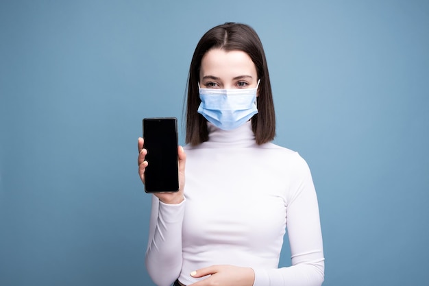 Uma jovem com uma máscara médica segura um telefone celular nas mãos e faz uma selfie em um fundo azul no estúdio.