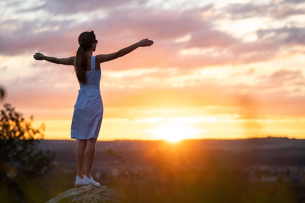 uma jovem com vestido de verão levantando as mãos em pé ao ar livre apreciando a vista