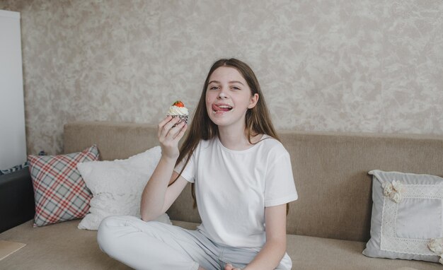 Uma jovem comendo um bolinho delicioso untado com creme sentada em casa no sofá