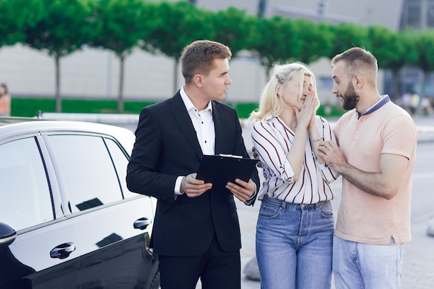 Uma jovem concessionária de terno mostra aos compradores um carro novo. Jovem casal, homem e mulher, compre um carro. Compra de máquinas, test drive.