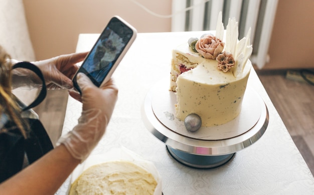Uma jovem confeiteira com um avental cinza tira fotos de um bolo preparado.