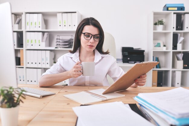 Uma jovem contadora de óculos, tomando chá ou café, segurando a prancheta com o documento à sua frente e lendo-o