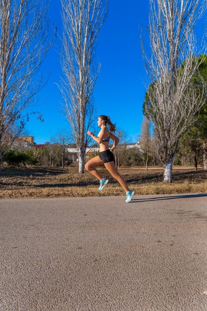 Uma jovem corredora vestida com calções apertados e um top branco correndo com passo longo e perna esticada com estilo e movimentos técnicos perfeitos em um caminho de asfalto em um parque natural